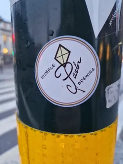 Street sticker A round sticker featuring the logo of Humble Parlor Brewing. The logo includes a stylized kite and the company's name. The words "Humble" and "Brewing" flank the logo. The sticker is affixed to a dark green pole with a yellow band at the bottom.