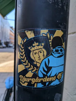 Street sticker The sticker is rectangular and predominantly features a stylized crest. The background has radiating yellow lines on a black base. On the left side of the crest, there is a golden laurel wreath. Within the crest, a golden outline surrounds a profile of a person's head wearing a crown. To the right of the crest, a figure in a bright blue jacket with a black cap and goggles is depicted. The text 'Djurgardens IF' is prominently displayed in a golden, stylized font across the bottom of the sticker.