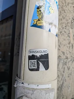 Street sticker A rectangular sticker with rounded corners, featuring a monochrome design. At the top, the text 'JHNSKGLND' is displayed in a sans-serif font. Below the text, there's a black and white landscape image depicting a winding path or boardwalk leading through a dense, dark forest or overgrown area. A prominent QR code is embedded on the left side of the path within the landscape image. The overall aesthetic is stark and somewhat mysterious.