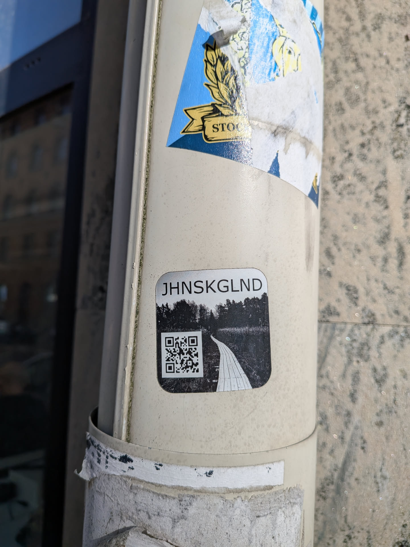 A rectangular sticker with rounded corners, featuring a monochrome design. At the top, the text 'JHNSKGLND' is displayed in a sans-serif font. Below the text, there's a black and white landscape image depicting a winding path or boardwalk leading through a dense, dark forest or overgrown area. A prominent QR code is embedded on the left side of the path within the landscape image. The overall aesthetic is stark and somewhat mysterious.