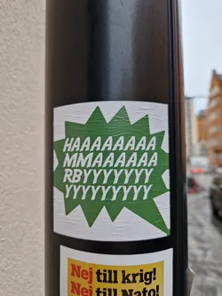 A square sticker with a green speech bubble shape containing the text "HAAAAAAA MMMMMMMMM RBYYYYYYYY YYYYYYYYYY" in white. The sticker is attached to a black pole, showing some peeling at the edges.