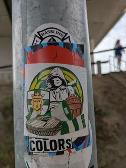 Street sticker The central sticker depicts a stylized hooded figure holding an old-fashioned brown football that has the word 'ULTRA' inscribed on it. To the left of the hooded figure, there is a shield with green and white vertical stripes, featuring a crowned head, possibly a queen or a historical royal figure. Below these elements, a modern football stadium is illustrated. The entire central design is encircled by a green and yellow sunburst or wreath pattern. The sticker predominantly uses shades of green, white, black, brown, and yellow/gold. Above and below this main sticker, fragments of other stickers are visible, bearing the words 'BASSLINE' and 'COLORS' respectively.