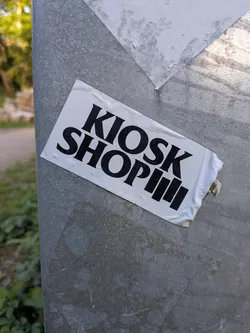 Street sticker A rectangular white sticker featuring bold black text 'KIOSK SHOP' followed by three vertical bars, affixed at an angle to a metallic post.