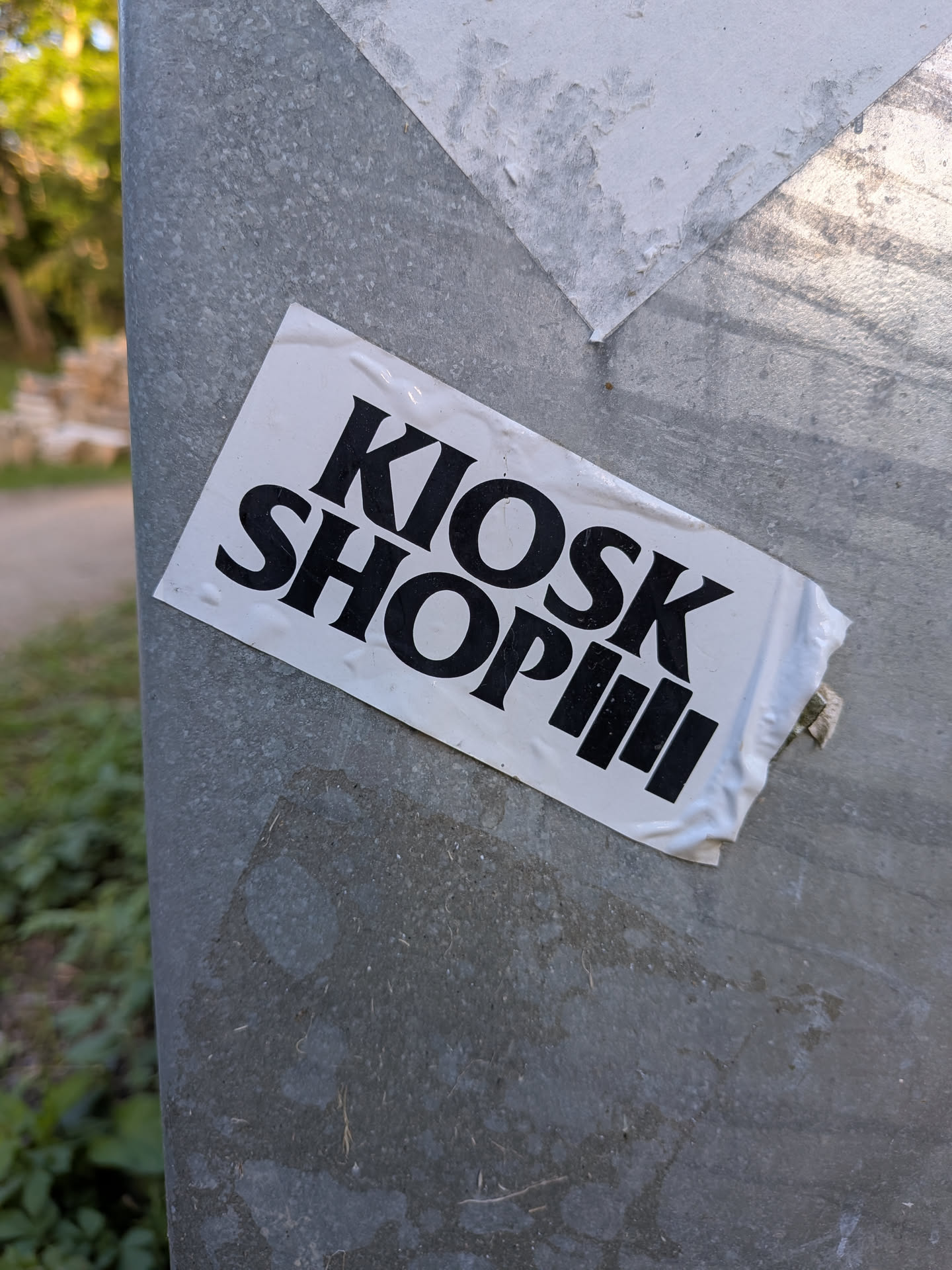 A rectangular white sticker featuring bold black text 'KIOSK SHOP' followed by three vertical bars, affixed at an angle to a metallic post.