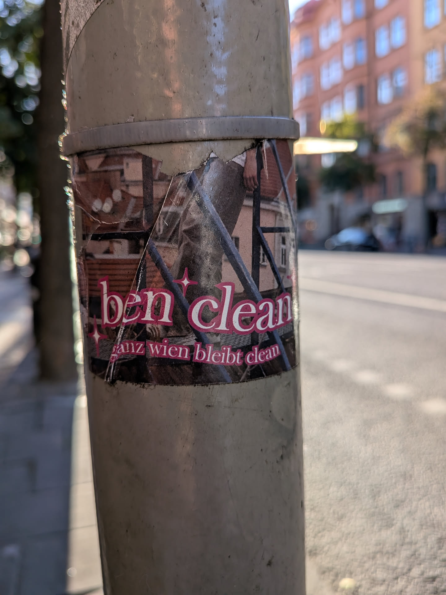 The sticker features bold, pink lettering that reads 'ben clean' with small star accents. Below this, in a smaller, also pink font, it says 'ganz wien bleibt clean'. The background of the sticker appears to be a collage of urban imagery, including what looks like a person's legs and a building facade with tiled roofs. The sticker is applied to a cylindrical pole and shows signs of wear and tear, with some peeling at the edges.