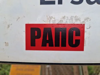 A rectangular sticker with a red background and black Cyrillic text. The text reads "РАПС". The sticker is affixed to a weathered white surface.