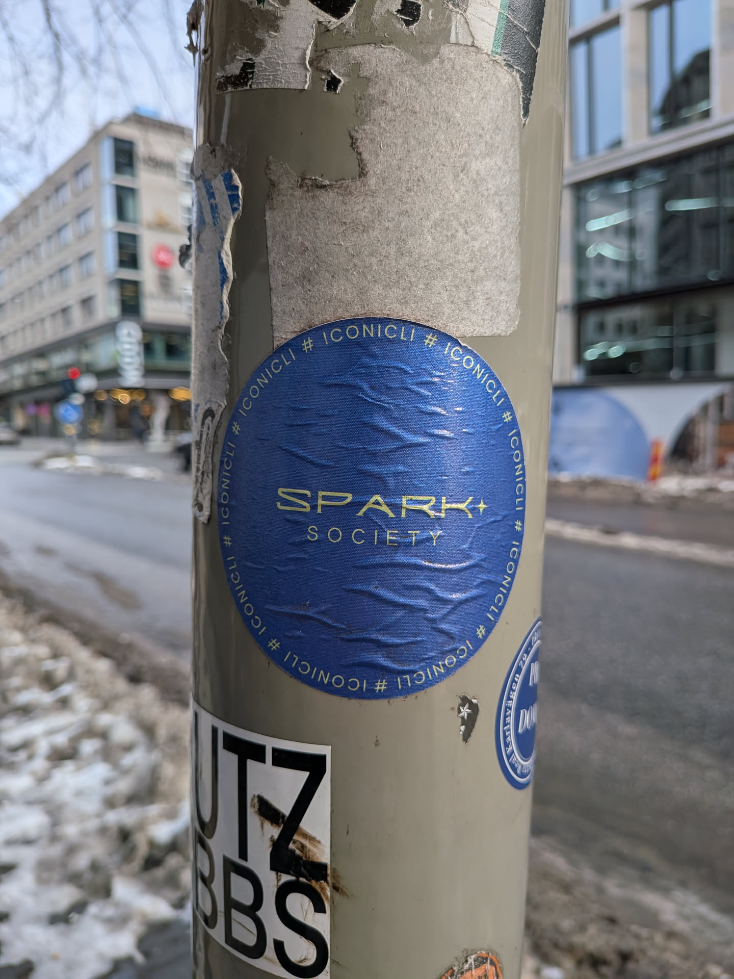 The sticker is circular, featuring a deep royal blue background with a visible textured, slightly wrinkled surface. In the center, the words "SPARK SOCIETY" are prominently displayed in a metallic gold or light yellow sans-serif font. "SPARK" is larger, with a small plus sign integrated into the 'K', positioned above the smaller word "SOCIETY". Encircling the central text, the hashtag "#ICONICLI" is repeated multiple times in the same gold/yellow color, following the curve of the sticker's edge. The sticker is affixed to a light grey pole and shows signs of wear and creasing.