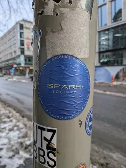 Street sticker The sticker is circular, featuring a deep royal blue background with a visible textured, slightly wrinkled surface. In the center, the words "SPARK SOCIETY" are prominently displayed in a metallic gold or light yellow sans-serif font. "SPARK" is larger, with a small plus sign integrated into the 'K', positioned above the smaller word "SOCIETY". Encircling the central text, the hashtag "#ICONICLI" is repeated multiple times in the same gold/yellow color, following the curve of the sticker's edge. The sticker is affixed to a light grey pole and shows signs of wear and creasing.