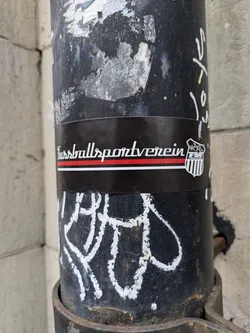 Street sticker A black and white sticker featuring the text "Fussballsportverein" in a stylized font. A small emblem is visible on the right.  The sticker is affixed to a dark metal post covered in other graffiti and markings.
