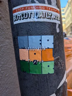 Street sticker The central sticker is square-shaped and features a geometric design composed of six rectangular blocks arranged in a 2x3 grid. The top row consists of two light blue/grey blocks, the middle row has two orange/peach blocks, and the bottom row has two green blocks. Thin black lines separate these colored sections. The right block in each row (blue, orange, green) contains a small black circular dot and has a jagged, 'M'-shaped edge on its right side. The left block in each row also has a jagged edge on its right side. Overlaid on this design are two lines of white text. The upper text, 'SAGOFRÖN', is in a clean, sans-serif font and spans across the upper part of the blue and orange sections. The lower text, 'Triptyk', is in a handwritten-style font and spans across the lower part of the orange and green sections. The overall aesthetic is modern and artistic with a muted color palette.