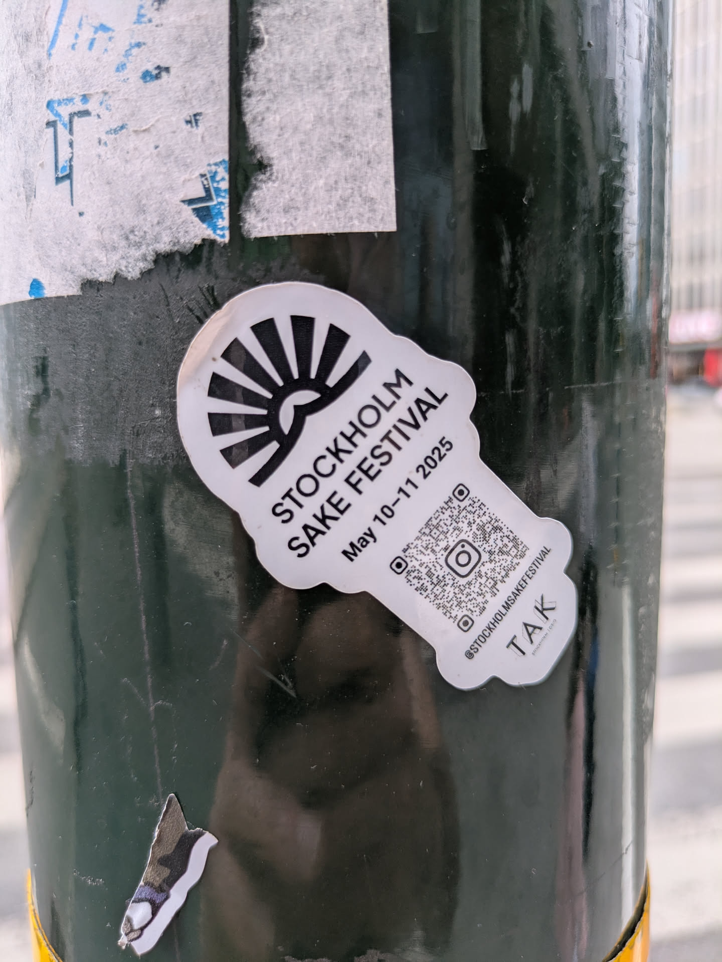 A distinctive black and white die-cut sticker shaped somewhat like a sake tokkuri (flask) with a rising sun motif at its top. The sticker advertises the 'Stockholm Sake Festival' scheduled for 'May 10-11 2025'. It prominently features a QR code with an embedded Instagram logo, alongside the handle '@STOCKHOLMSAKEFESTIVAL'. Below the main information, the text 'TAK' and 'STOCKHOLM | OSLO' are visible. The sticker is affixed to a dark green, possibly metallic, pole, with older, torn paper remnants visible around it.