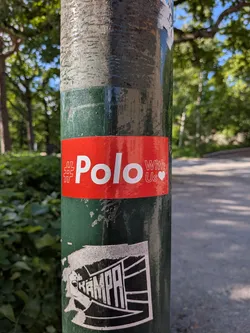 Street sticker A rectangular red sticker on a green pole featuring white text that reads '#Polo With Us' next to a small white heart.