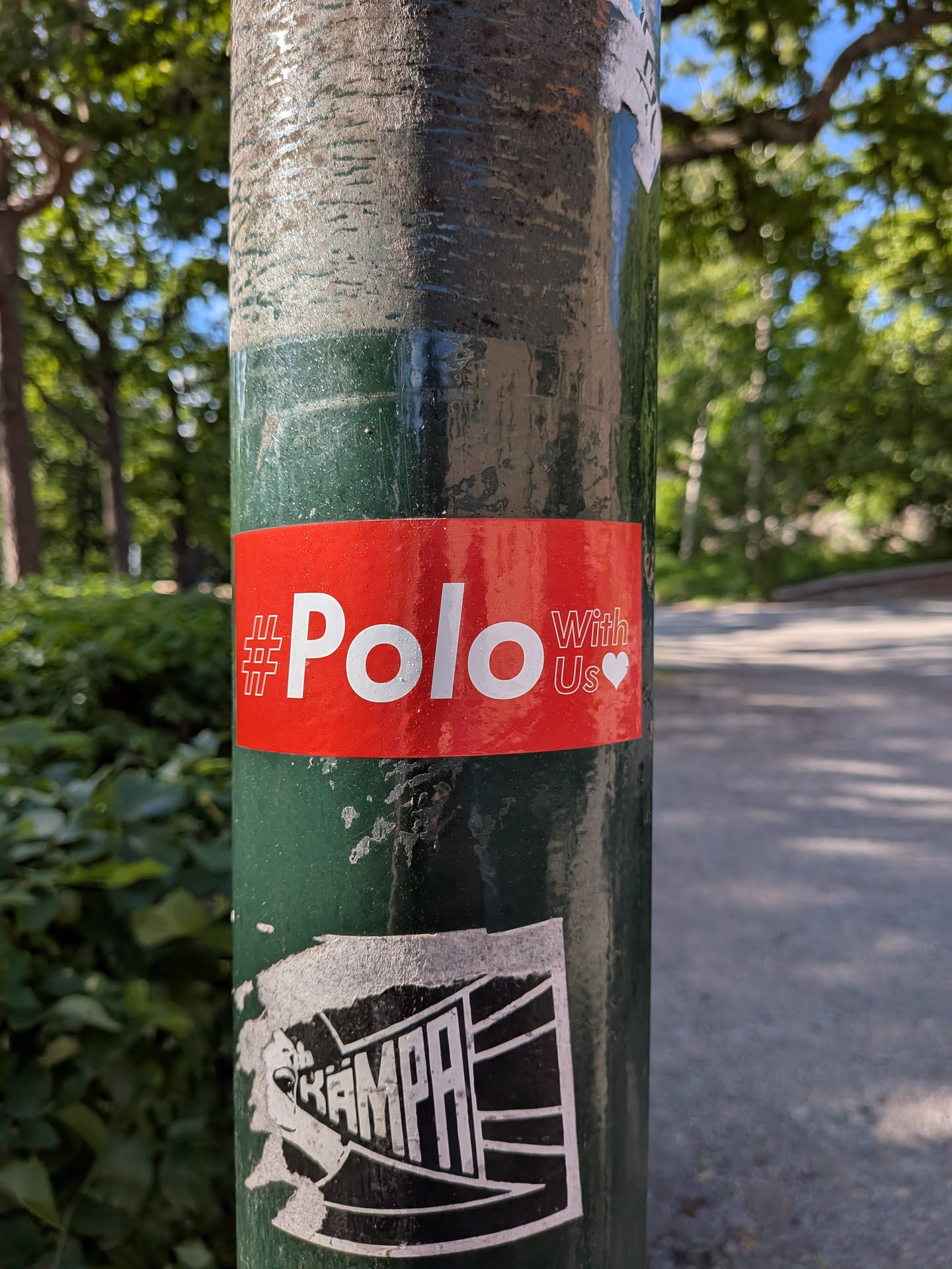 A rectangular red sticker on a green pole featuring white text that reads '#Polo With Us' next to a small white heart.
