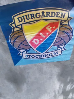Street sticker A sticker featuring the logo of Djurgården D.I.F., a sports club from Stockholm, Sweden. The logo is a shield with the club's initials and name, flanked by two American footballs. The colors are yellow, pink, blue, and white. The sticker is attached to a metal surface.