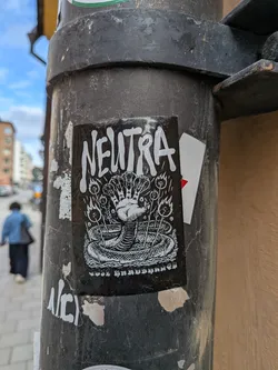Street sticker The sticker is a black and white illustration. At the top, the word "NEUTRA" is prominently displayed in a distressed, bold, uppercase font. Below the text, the central image features a coiled snake at the bottom, its body intricately textured with scales. Emerging from the center of the snake's coil is a human-like hand, but it appears to have multiple fingers or digits, resembling a cluster of hands or a hydra-like appendage. Surrounding the hand and snake are several stylized plants or stalks, each topped with a small flame or burning element, emitting small dots or sparks. The overall aesthetic is dark, intricate, and somewhat mystical or occult, rendered in a stark black and white contrast.