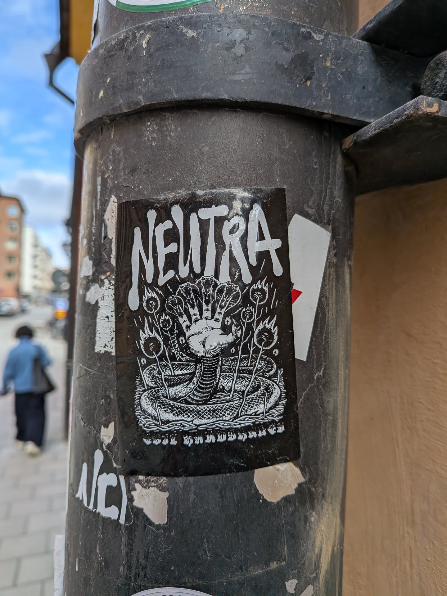 The sticker is a black and white illustration. At the top, the word "NEUTRA" is prominently displayed in a distressed, bold, uppercase font. Below the text, the central image features a coiled snake at the bottom, its body intricately textured with scales. Emerging from the center of the snake's coil is a human-like hand, but it appears to have multiple fingers or digits, resembling a cluster of hands or a hydra-like appendage. Surrounding the hand and snake are several stylized plants or stalks, each topped with a small flame or burning element, emitting small dots or sparks. The overall aesthetic is dark, intricate, and somewhat mystical or occult, rendered in a stark black and white contrast.