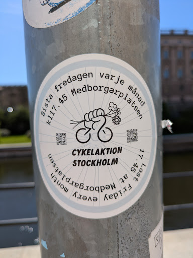 This circular sticker promotes 'Cykelaktion Stockholm', a cycling event held on the last Friday of every month at Medborgarplatsen at 17:45. The sticker features an illustration of a fist holding flowers with a bicycle wheel design.