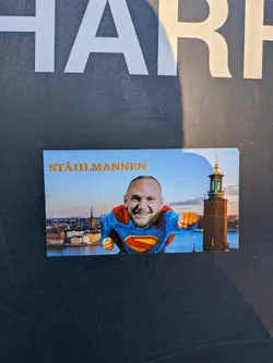 Street sticker The sticker features a humorous image of a bald, bearded man's smiling face photoshopped onto the body of Superman, complete with the classic blue suit, red cape, and yellow and red 'S' emblem. He is depicted in a flying pose with a clenched fist. The background is a scenic panoramic view of Stockholm, Sweden, showcasing its waterfront buildings, spires, and the distinctive tower of the Stockholm City Hall (Stadshuset) on the right. The sky is a bright blue with a soft gradient. In the upper left corner, the text "STÅHLMANNEN" is written in golden, embossed-style capital letters.