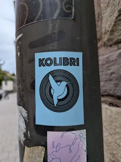 Street sticker A rectangular sticker with a light blue background, adhered to a dark metal pole. At the top, the word 'KOLIBRI' is printed in a bold, black, sans-serif font. Below the text is a circular graphic resembling a vinyl record with concentric black rings. In the center of this circle, a silhouette of a hummingbird in flight is depicted in light blue, set against a solid black circular background.
