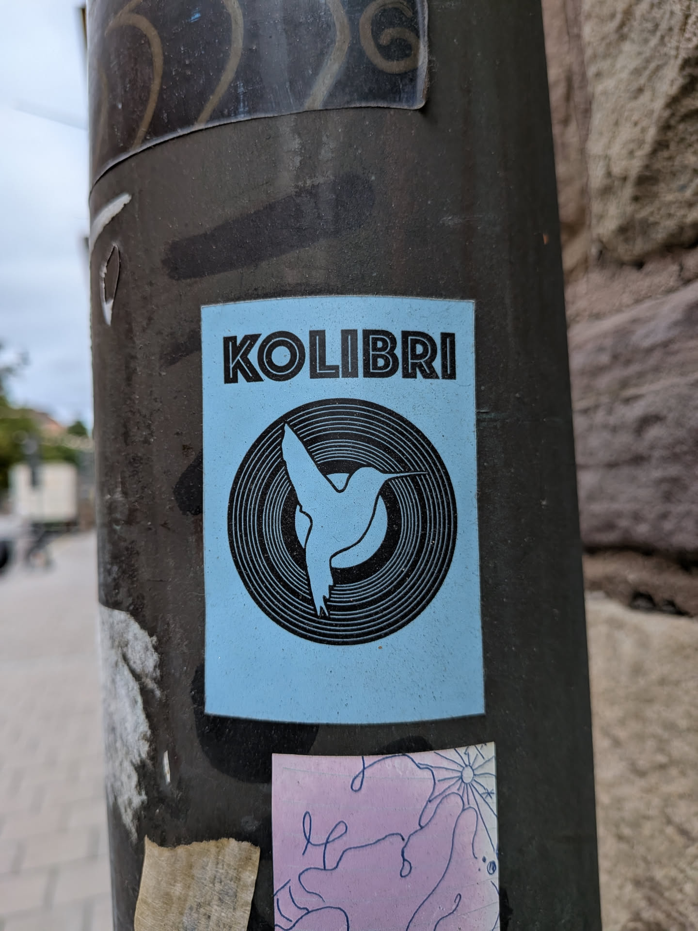 A rectangular sticker with a light blue background, adhered to a dark metal pole. At the top, the word 'KOLIBRI' is printed in a bold, black, sans-serif font. Below the text is a circular graphic resembling a vinyl record with concentric black rings. In the center of this circle, a silhouette of a hummingbird in flight is depicted in light blue, set against a solid black circular background.