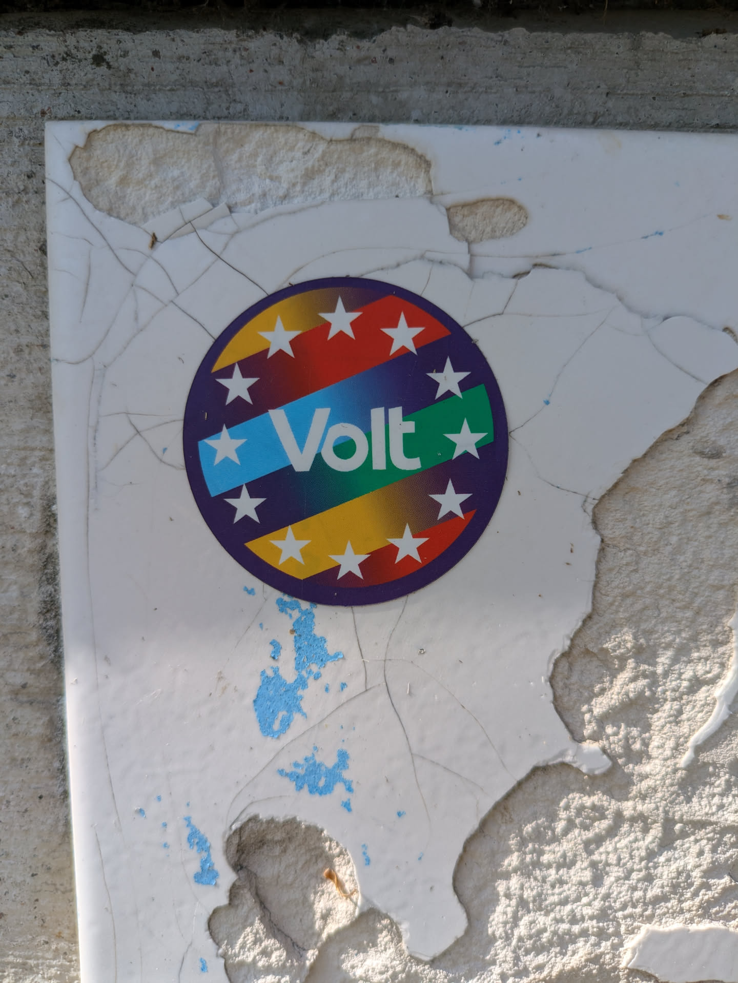 This sticker for Volt is a colorful round emblem with stars around the edge and the word "Volt" written across a blue, green and yellow stripe. The sticker is adhered to a damaged and cracked surface, which adds to the sticker's weathered appearance.