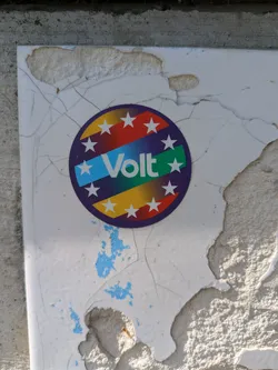 Street sticker This sticker for Volt is a colorful round emblem with stars around the edge and the word "Volt" written across a blue, green and yellow stripe. The sticker is adhered to a damaged and cracked surface, which adds to the sticker's weathered appearance.