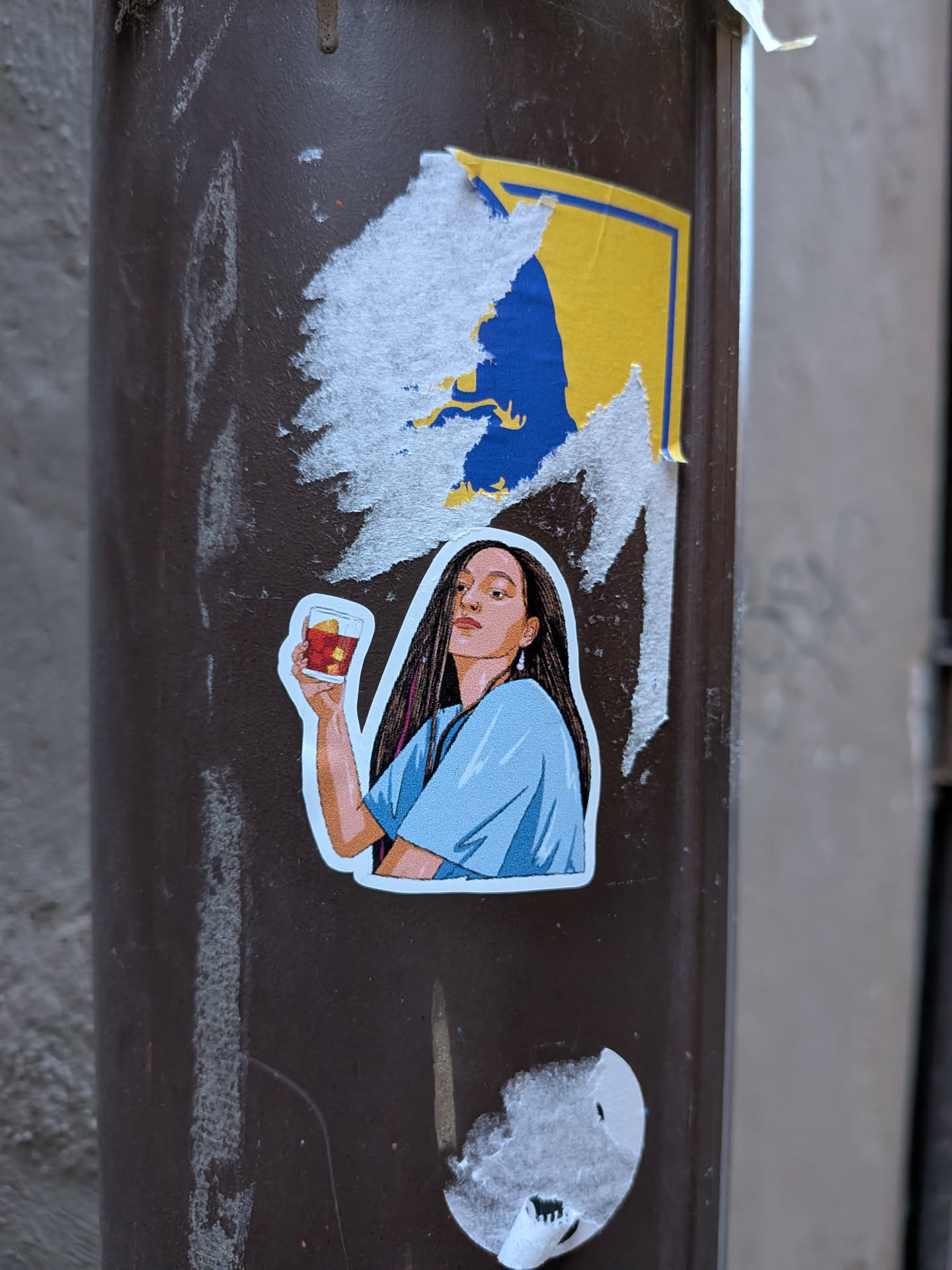 Street sticker The sticker features an illustration of a young woman from the chest up, looking slightly upwards and to her right with a confident expression. She has long, dark, straight hair with subtle pinkish-purple streaks visible on the left side. Her skin tone is warm. She is wearing a light blue, short-sleeved top with a V-neck. Her right hand (viewer's left) is raised, holding a clear glass containing a reddish-brown drink with ice cubes and a yellow garnish (possibly a lemon peel or fruit). The art style is clean, with distinct lines and flat colors, giving it a modern graphic novel or cartoon aesthetic. The sticker has a clean white border around the illustration and is placed on a dark brown pole, with remnants of a torn yellow and blue sticker above it.