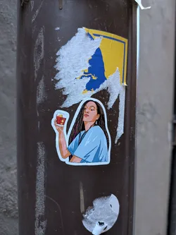 Street sticker The sticker features an illustration of a young woman from the chest up, looking slightly upwards and to her right with a confident expression. She has long, dark, straight hair with subtle pinkish-purple streaks visible on the left side. Her skin tone is warm. She is wearing a light blue, short-sleeved top with a V-neck. Her right hand (viewer's left) is raised, holding a clear glass containing a reddish-brown drink with ice cubes and a yellow garnish (possibly a lemon peel or fruit). The art style is clean, with distinct lines and flat colors, giving it a modern graphic novel or cartoon aesthetic. The sticker has a clean white border around the illustration and is placed on a dark brown pole, with remnants of a torn yellow and blue sticker above it.