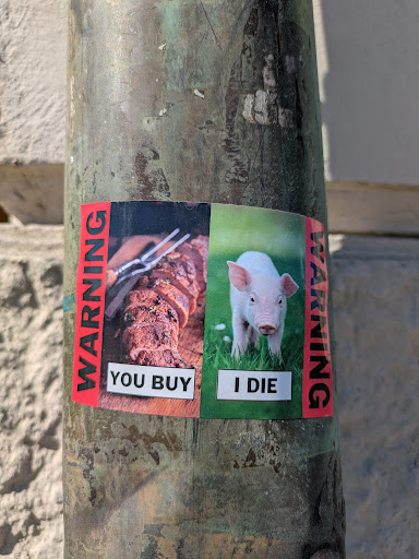 The sticker is divided into two sections. The left side depicts a cooked meat dish, alongside the words "YOU BUY." The right side shows a piglet in a grassy field, accompanied by the words "I DIE." A red warning label is placed vertically on each side of the sticker.