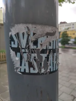 Street sticker A weathered sticker affixed to a gray pole.  Much of the sticker is torn and peeling, obscuring some of the text. The visible text appears to read, "SLEEPING JASTARE", in a sans-serif font, with light-blue lettering on a black background. The sticker shows significant wear and tear.