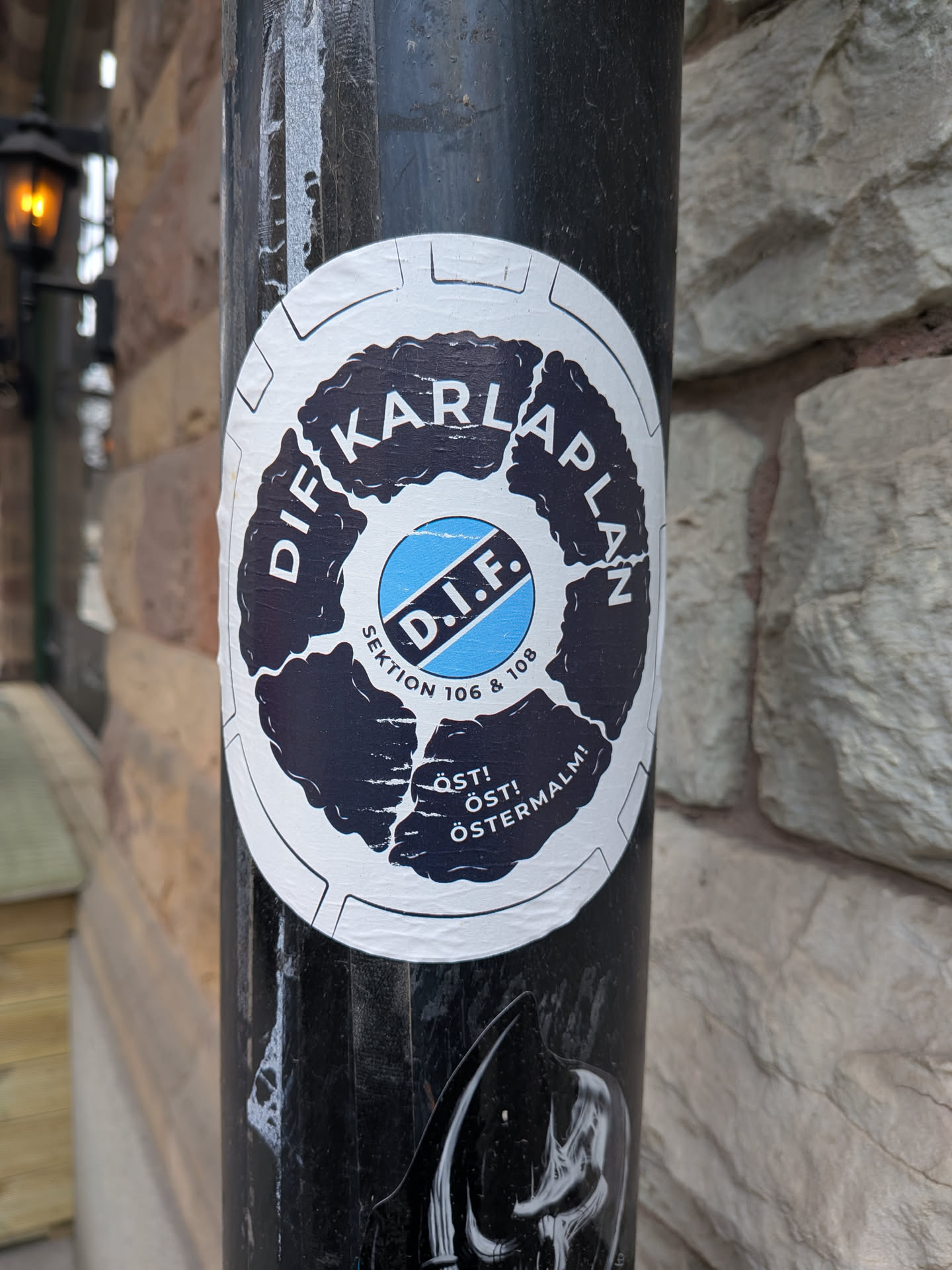 The sticker is circular with a white background and a prominent black, distressed, gear-like border. Inside this border, the words 'DIF KARLAPLAN' are written in white capital letters, following the curve of the circle. The center features a light blue circle with a black diagonal stripe, containing the white capital letters 'D.I.F.'. Below this central emblem, in a smaller font, is 'SEKTION 106 & 108'. At the bottom, also in white capital letters, are the words 'ÖST! ÖST! ÖSTERMALM!', with the 'ÖST!' repeated twice. The overall design uses a limited palette of white, black, and light blue, with a rugged, somewhat worn aesthetic.