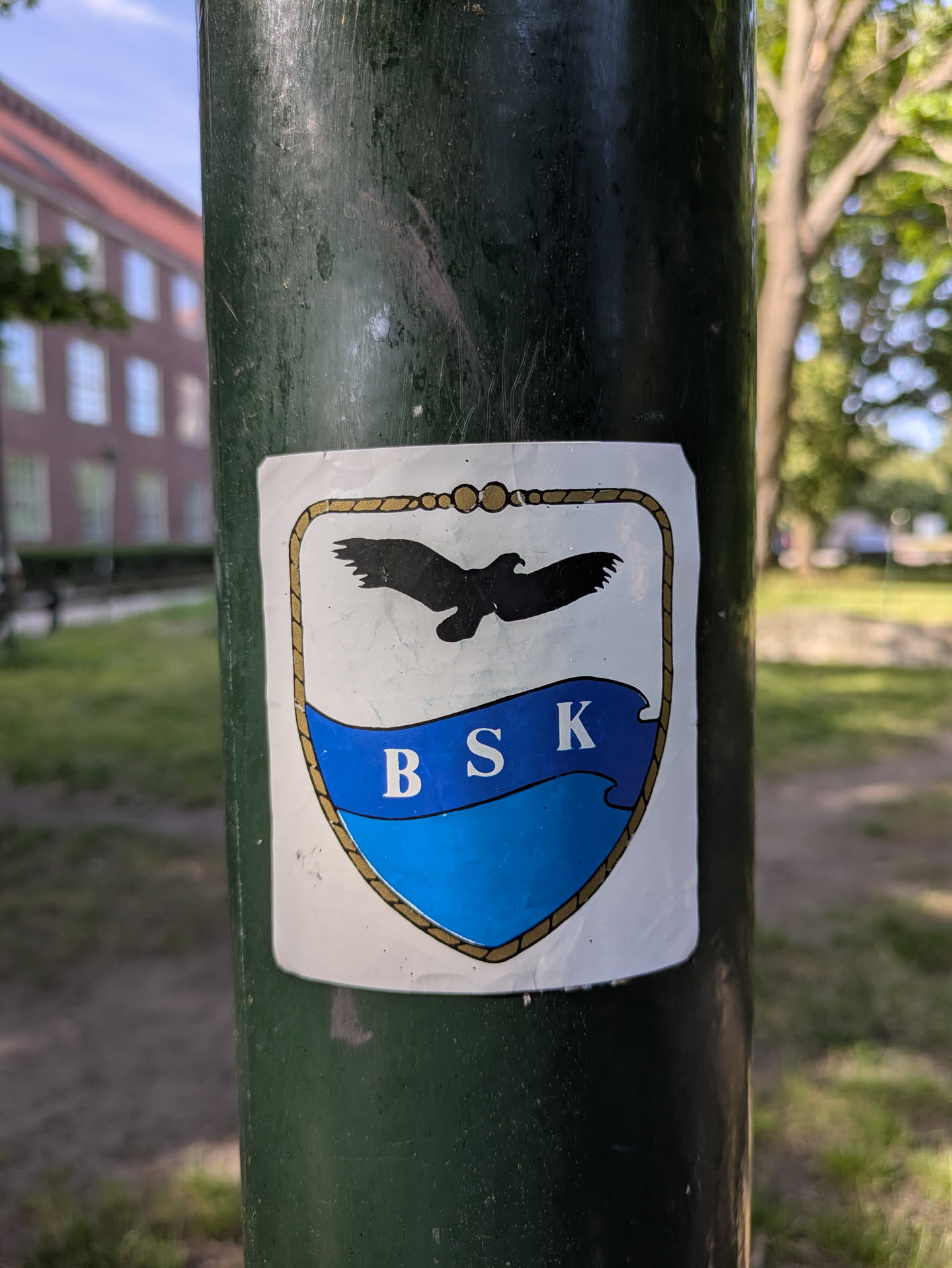 A weathered sticker on a dark green pole featuring a crest-like design. The sticker has a white background with a gold rope border, a black silhouette of an eagle flying above a blue wave banner, and the letters "BSK" in white on the blue banner.