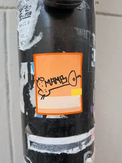 Street sticker The sticker is a rectangular label with slightly rounded corners, featuring a vibrant orange background that subtly fades to a lighter peach tone towards the bottom. A thin, darker orange border frames the sticker. At its center is a black, hand-drawn doodle depicting a small, rounded creature, possibly a mouse or rat, with two dots for eyes and whiskers. A thick black line extends from this creature to an elongated oval shape with a small circle inside, resembling a simple bell or abstract form. Above this line, the word 'MAMA' is written in bold, blocky, hand-drawn black letters. On the right edge of the sticker, there's a small yellow rectangular strip and a vertical white strip with the text 'W11003' printed in tiny red characters. The sticker is affixed to a textured black pole, surrounded by remnants of other torn and faded stickers.