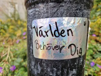 Street sticker The sticker is oval-shaped with a holographic, iridescent finish that reflects light in shades of blue, pink, and yellow. It is applied to a textured, dark gray surface.  The text is handwritten in black, cursive script. The first line reads 'Världen' (The world) and the second line reads 'Behöver Dig' (needs you). The overall appearance is eye-catching due to the holographic material and the personal, handwritten text.