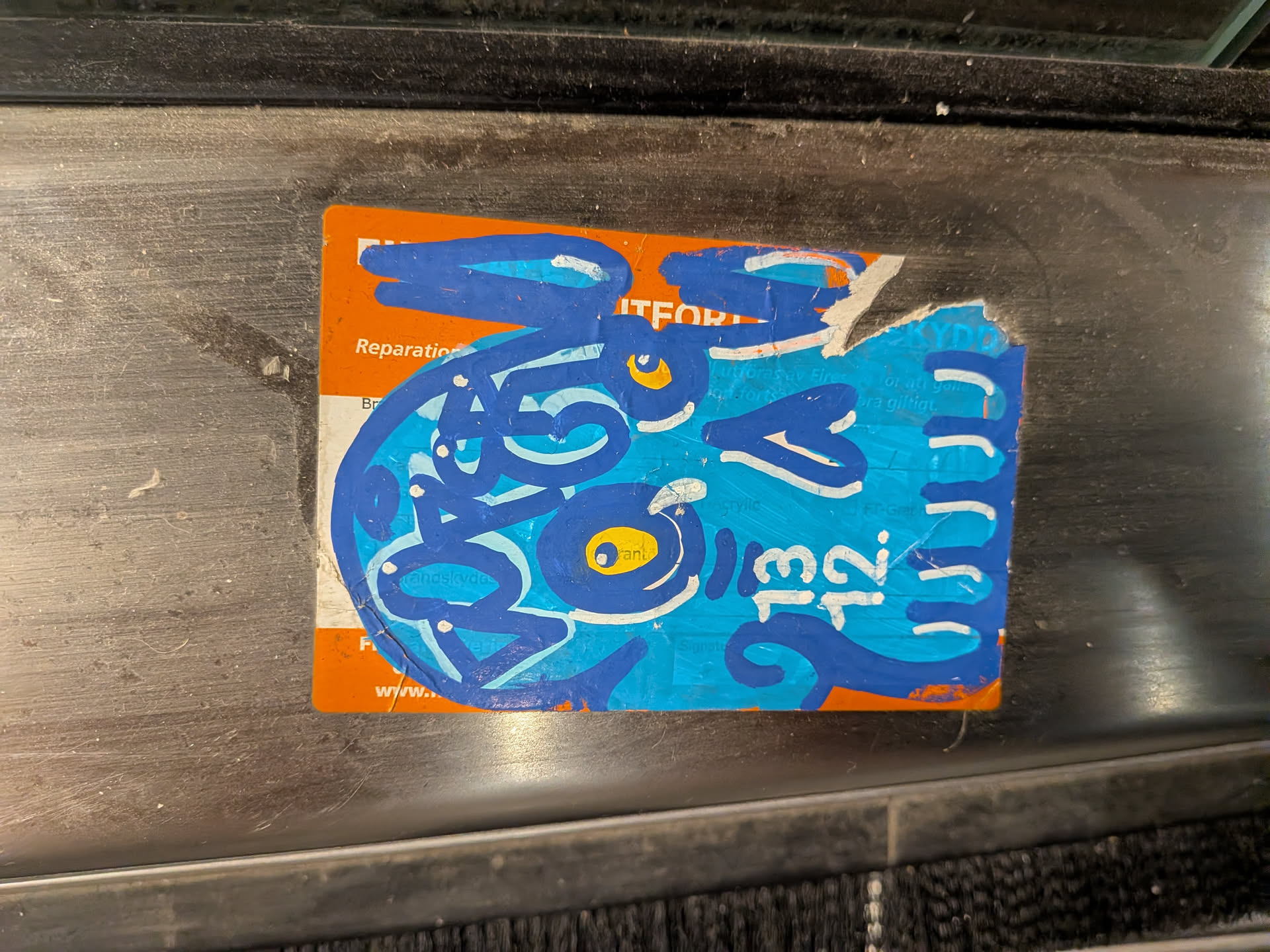 The sticker is a rectangular piece, likely a repurposed label or card, with torn and irregular edges, particularly on the right side. It is affixed to a brushed metal surface. The base layer of the sticker is an orange color, over which a stylized, abstract creature or character has been painted in various shades of blue, accented with yellow and white. The creature features two prominent yellow eyes with dark blue pupils. Its body is composed of thick, swirling dark blue lines, giving it a dynamic, graffiti-like appearance. Lighter blue and white strokes add texture and highlight to the design. On the right side, partially integrated into the blue design, are the white numbers '13' and '12' stacked vertically. Faint text from the original orange label is visible underneath the blue paint in several areas.