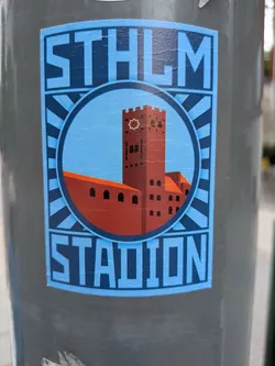 Street sticker Rectangular sticker depicting the red brick Stockholm Stadion tower inside a circular frame. Features a blue sunburst background and bold text reading 'STHLM' and 'STADION'.