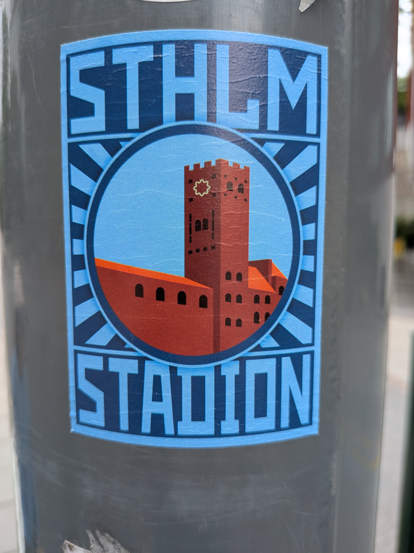 Rectangular sticker depicting the red brick Stockholm Stadion tower inside a circular frame. Features a blue sunburst background and bold text reading 'STHLM' and 'STADION'.