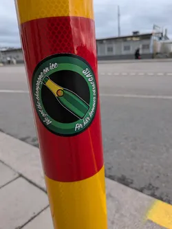 Street sticker The sticker is circular with a green border. The center features a stylized illustration of a green champagne bottle tipped diagonally, with its neck and cork pointing towards the upper left. The bottle has a yellow cap and a green label with a gold circular design on it. The text around the border reads 'We got the champagne on ice' and 'För här kommer vårat Gais'. The background of the circle is black.