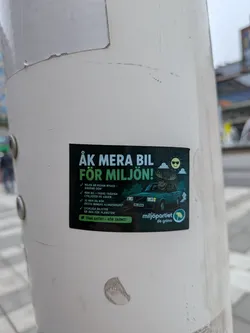 Street sticker A rectangular sticker with a dark green background. It features a cartoon moose wearing sunglasses driving a green Volvo 240 sedan. In the background, a smiling sun with sunglasses shines behind clouds. The text is written in Swedish, presenting satirical arguments for why driving is good for the environment, mimicking the branding of the Swedish Green Party ('Miljöpartiet de gröna') with their dandelion logo in the bottom right corner.