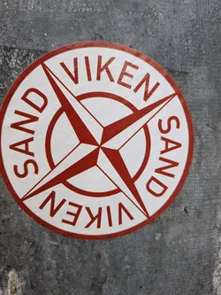 A circular sticker featuring a prominent red compass rose design on a white background. The compass rose has four main points and four smaller intermediate points, all radiating from a central red circle. Along the top curve of the sticker, the text 'SAND VIKEN' is written in bold red capital letters. The same text, 'VIKEN SAND', is repeated along the bottom curve, inverted relative to the top text. The sticker has a thin red border and appears slightly worn and textured, adhered to a rough, dark grey surface.