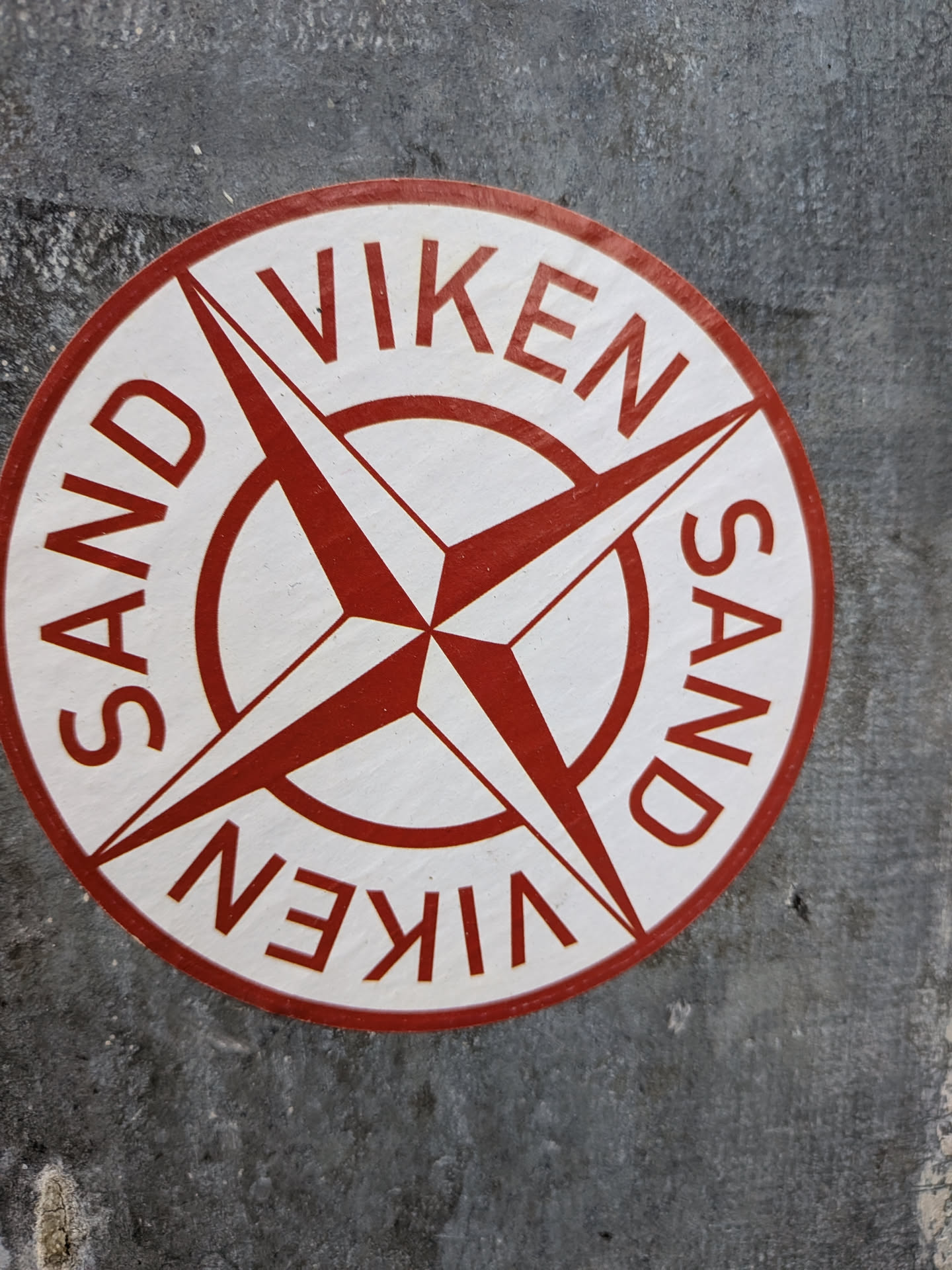 A circular sticker featuring a prominent red compass rose design on a white background. The compass rose has four main points and four smaller intermediate points, all radiating from a central red circle. Along the top curve of the sticker, the text 'SAND VIKEN' is written in bold red capital letters. The same text, 'VIKEN SAND', is repeated along the bottom curve, inverted relative to the top text. The sticker has a thin red border and appears slightly worn and textured, adhered to a rough, dark grey surface.