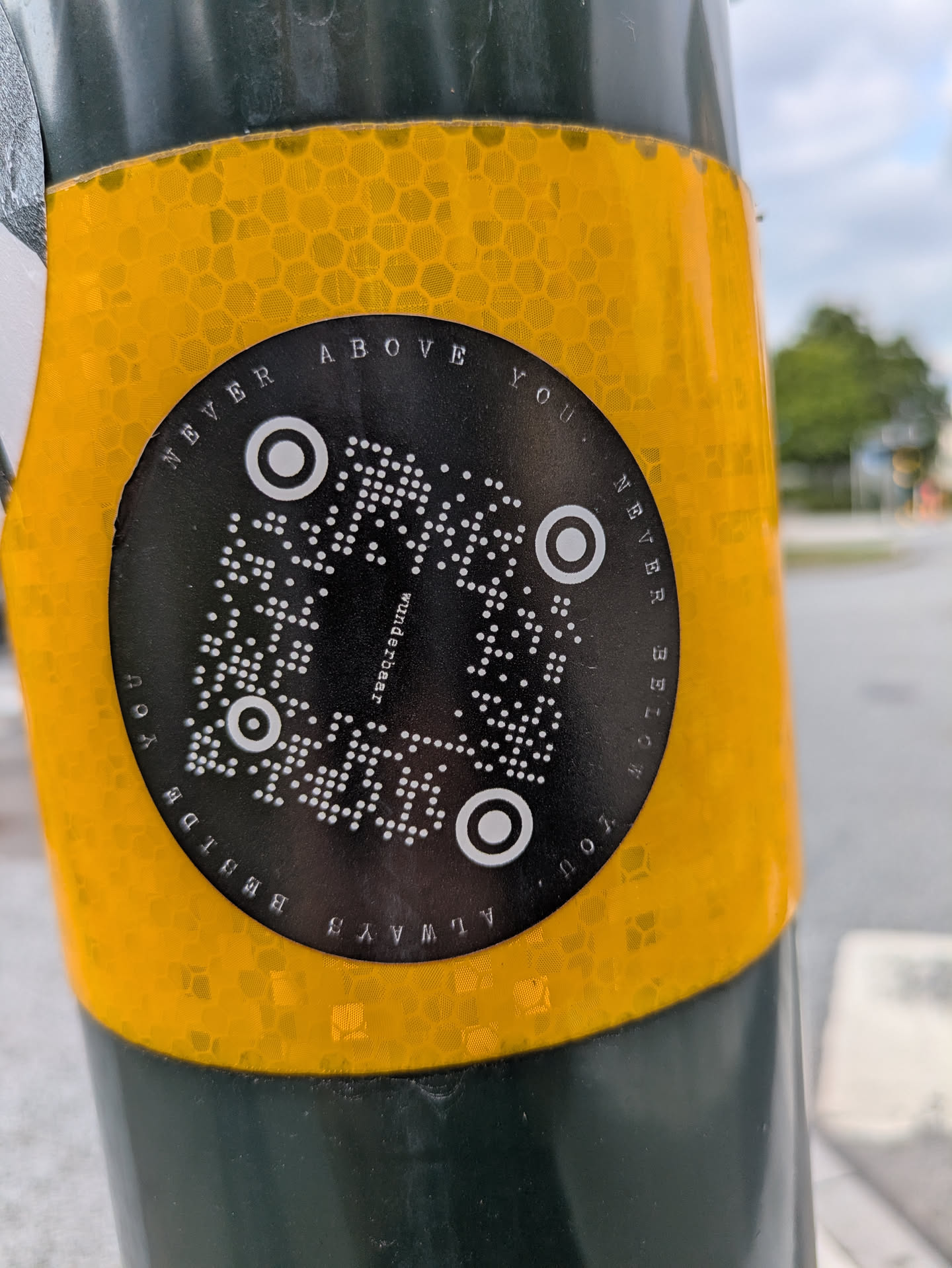 The sticker is circular, primarily black with white elements, adhered to a pole wrapped in a bright yellow, hexagonal-patterned reflective material. At its center is a QR code composed of white dots against the black background. Around the perimeter of the circle, white text is arranged in an arc, both at the top and bottom. The word 'wunderbear' is vertically integrated into the design of the QR code itself.