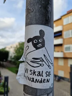 Street sticker Black and white illustration of a dog-like creature with a black mask on its face. The text at the bottom says "Vi skal se hinanden" which translates from Danish to English as "We shall see each other". The sticker is attached to a gray pole.