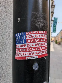 Street sticker A rectangular sticker designed like the US flag. The blue canton features white bomb silhouettes instead of stars. The red stripes contain the Swedish text 'DCA-AVTALET' and 'RIV UPP DCA-AVTALET'. A small white label at the bottom reads '★ UNG VÄNSTER'.
