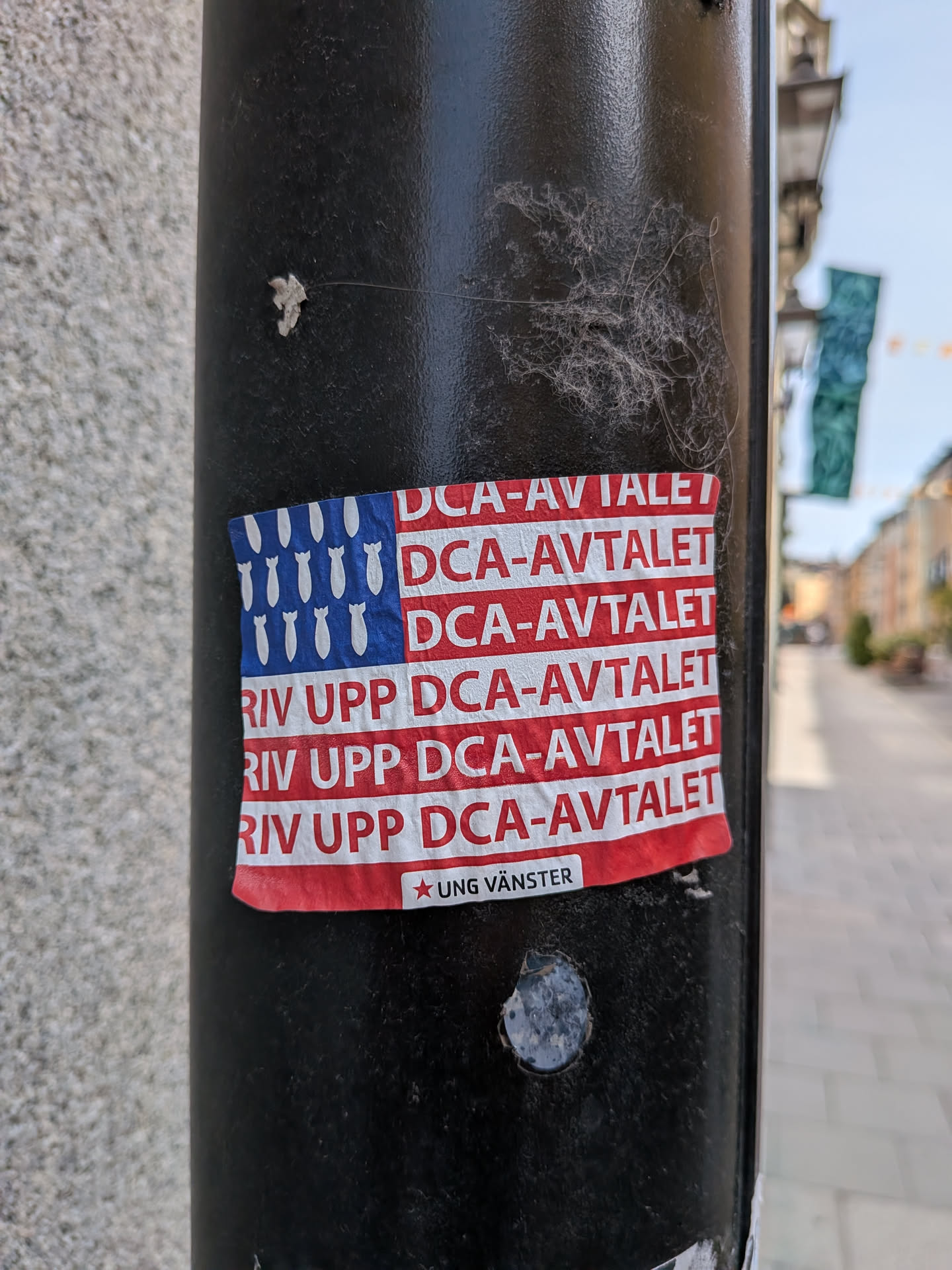 A rectangular sticker designed like the US flag. The blue canton features white bomb silhouettes instead of stars. The red stripes contain the Swedish text 'DCA-AVTALET' and 'RIV UPP DCA-AVTALET'. A small white label at the bottom reads '★ UNG VÄNSTER'.