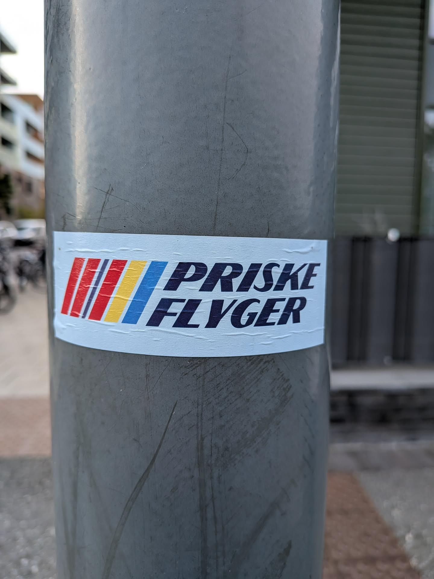 The sticker is rectangular with a light blue background. On the left side, there are four vertical stripes: red, dark red, yellow, and blue. To the right of the stripes, in large, bold, dark blue sans-serif font, are the words "PRISKE FLYGER". The sticker appears to be applied to a metal pole, and there are some minor wrinkles and creases on its surface.