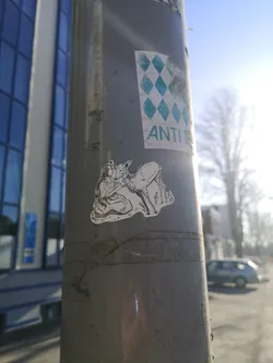 Street sticker The sticker features a monochromatic illustration of a woman with long, flowing hair gently embracing a deer. The woman is depicted in a seated or kneeling position, with her arm around the deer's neck, and their faces are close, suggesting a kiss or nuzzle. The deer stands beside her, looking towards her. Both figures are drawn with fine black lines on a white background, creating a detailed, illustrative style with some subtle shading. The sticker has an irregular, die-cut shape that follows the contours of the figures.