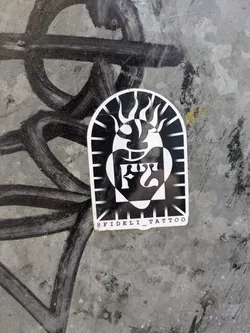 A white sticker with a black abstract image in the center, resembling a stylized sun or head. The sticker is arch-shaped at the top and has the text "@FIDELI_TATTOO" printed at the bottom. The sticker is stuck on a metallic surface that is marked with black paint.