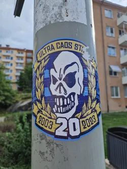Street sticker A sticker featuring a skull graphic with text that reads "ULTRA CAOS STAND 2003 20 2023". The skull is encircled by a laurel wreath-like design. The color scheme is primarily blue, yellow, and white.