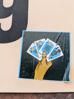 Street sticker A sticker featuring a hand holding three playing cards. Each card depicts a different football player in a light blue and black uniform. The style is illustrated and cartoonish, with a dark background and a light blue border around the sticker.